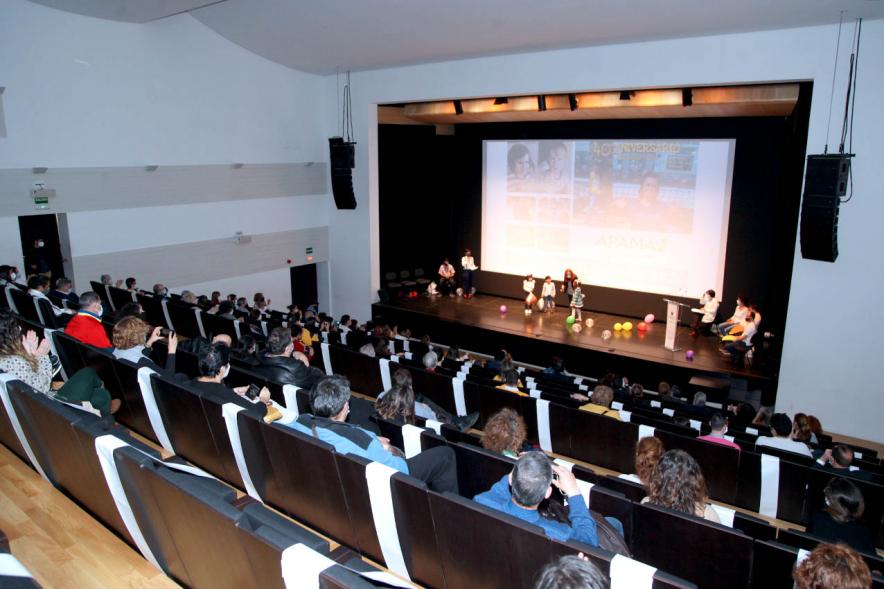 Presentación Calendario APAMA 2021 - Imagen panorámica, vemos el escenario donde vemos actuando a unas personas y al público asistente.