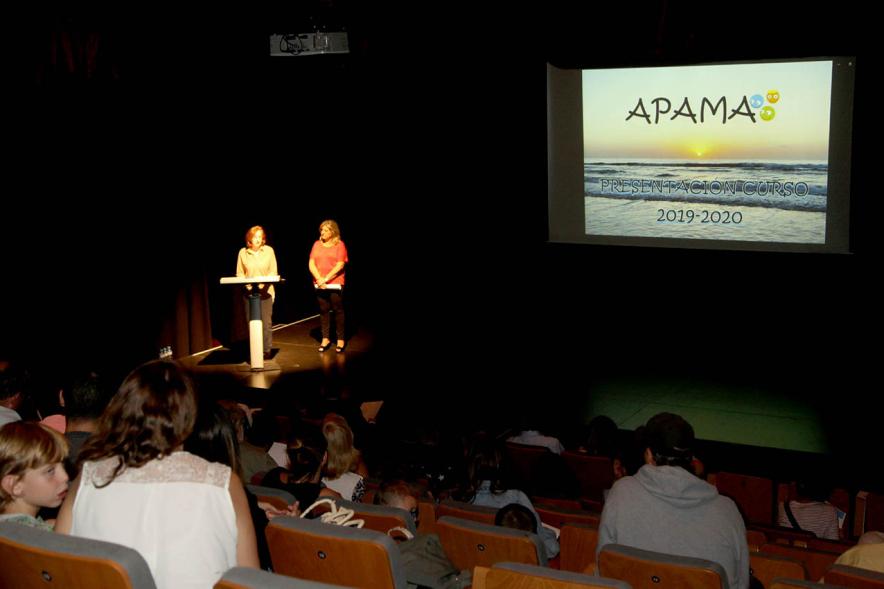 Presentación Curso Escolar 2019-20 APAMA - Panorámica del escenario, dos representantes y el cartel de la asociación.