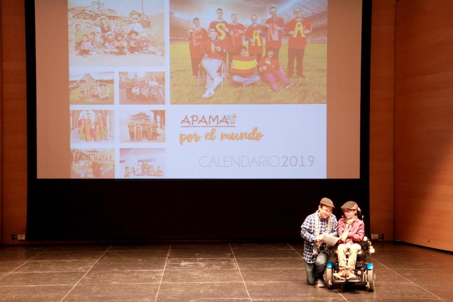 Presentación Calendario APAMA 2019 - Plano del escenario con un niño en silla de ruedas y su padre, detrás el cartel de la asociación iluminado.