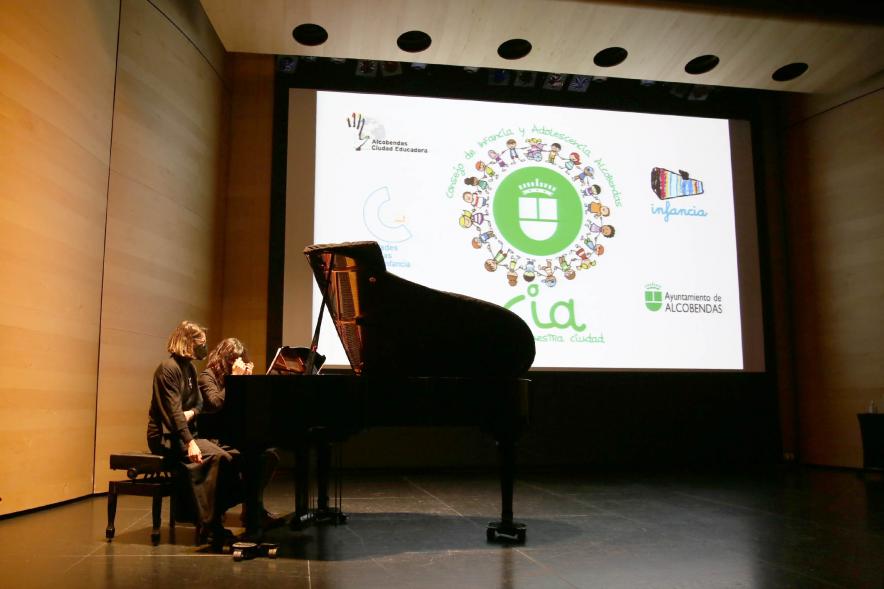 Nuevo Consejo de la Infancia y Adolescencia - Imagen de dos jóvenes al piano,  la pantalla con  el cartel del acto.