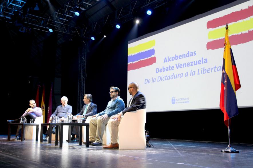 Alcobendas Debate Venezuela - Imagen de los ponentes en el escenario y el cartel del debate iluminado.