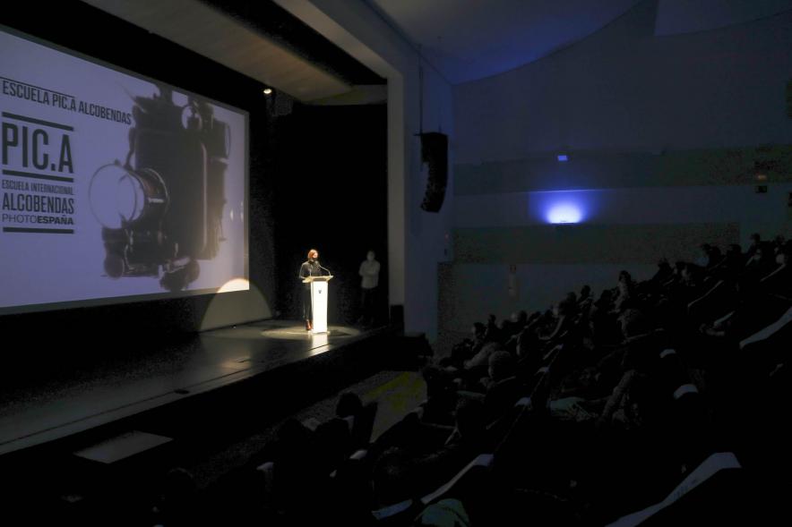 Entrega Diplomas PIC.A Curso 2019-20 - Imagen en penumbra del auditorio, solo iluminado el escenario con una autoridad municipal en el atril.