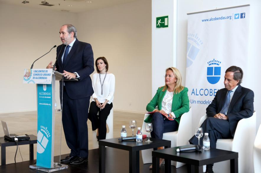 Ier Encuentro de Comercio Urbano de Alcobendas - Imagen del escenario donde un orador esta dirigiéndose a los asistentes al encuentro y los demás oradores.