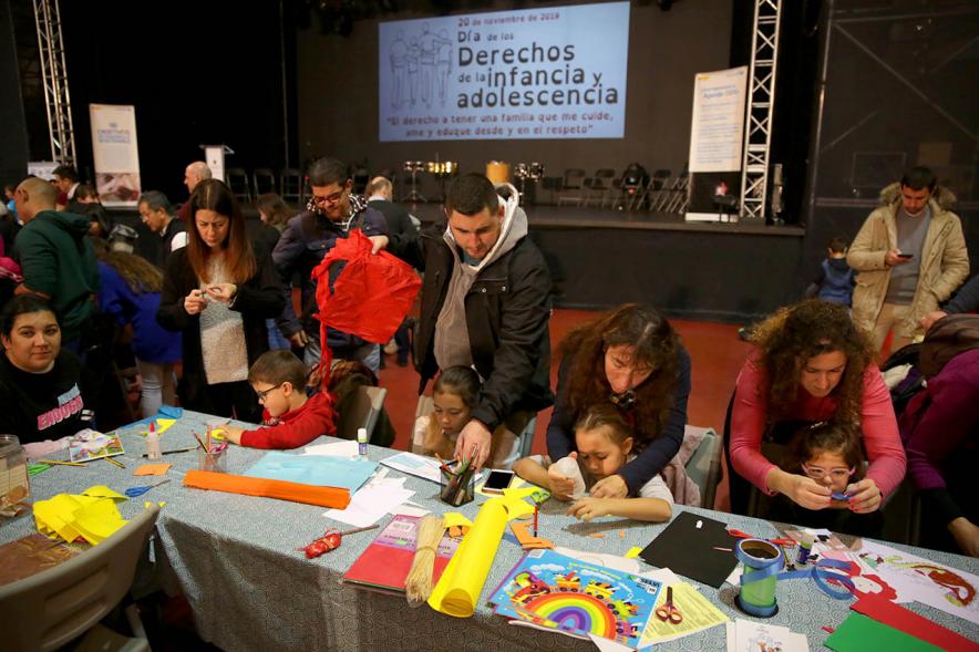 Fiesta Derechos de la Infancia -Primer plano de un grupo de niños con sus padres jugando y el cartel del evento en la pantalla.