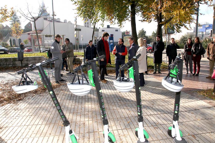 Presentación Patinetes Eléctricos Empresa LIme España -  Imagen en primer plano tenemos unos patinetes aparcados en la calle y al fondo vemos a las autoridades asistentes al acto.