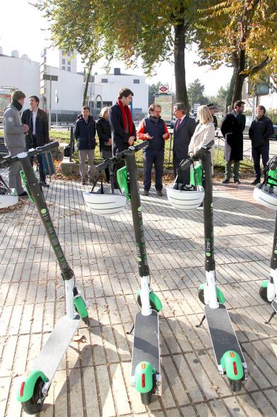 Presentación Patinetes Eléctricos Empresa LIme España - Imagen en primer plano tenemos unos patinetes aparcados en la calle y al fondo vemos a las autoridades asistentes al acto.