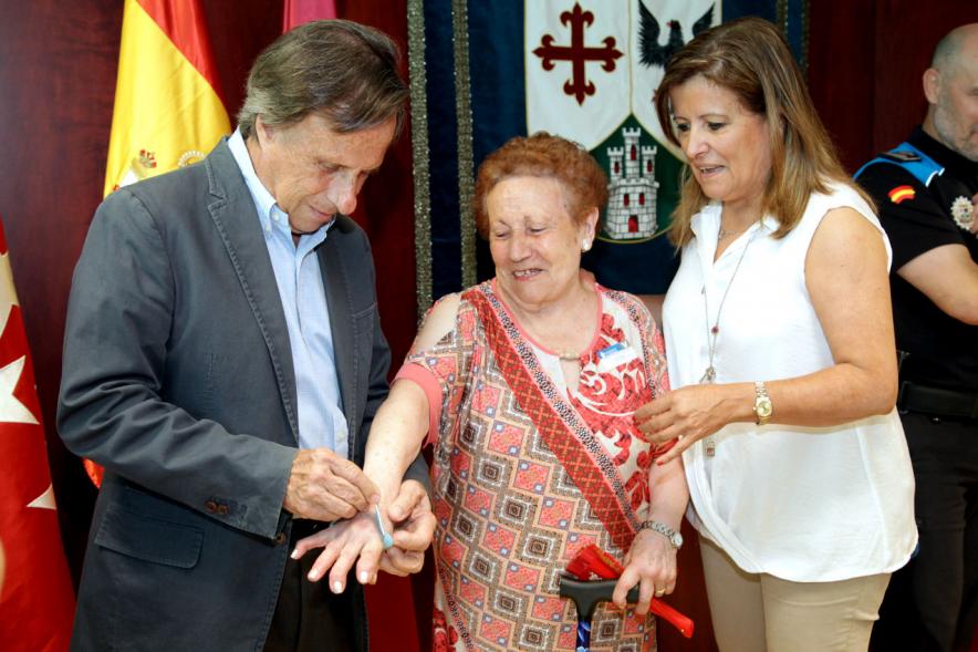 Entrega de Pulseras QR a la Asociación del Alzheimer - Imagen del alcalde García de Vinuesa colocando la pulsera en su muñeca a una mujer en presencia de otras autoridades.