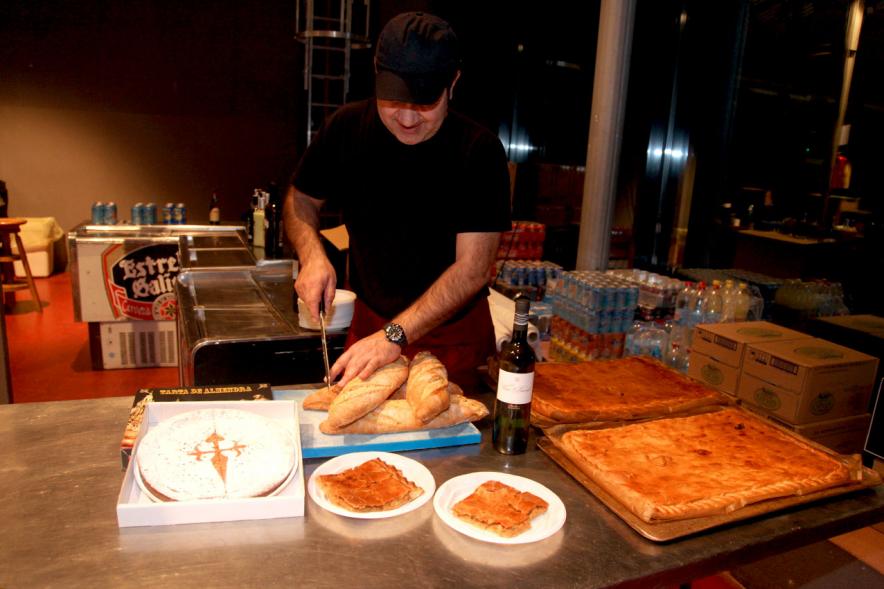 Semana Cultural Xuntanza de Galegos - Inauguración de la Romeria del Pulpo y el Mejillón - Imagen de persona cortando pan y platos de empanada gallega.