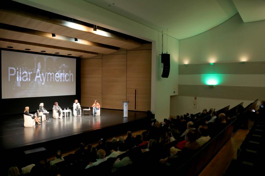  Coloquio y Visita Guiada a la Exposición "Cinco Miradas"  - Imagen vista panorámica lejana del escenario donde vemos a las participantes en el coloquio.