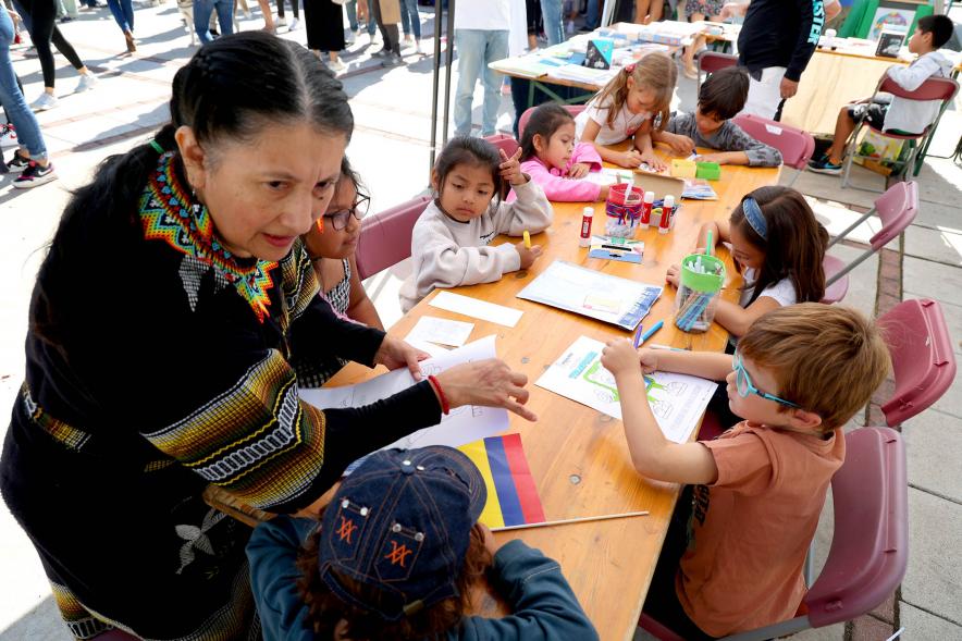 Festival Intercultural Mercado Gastronómico y Artesanía Día de la Hispanidad -  Primer plano de una mesa de actividades con la coordinadora con el traje de su pais.